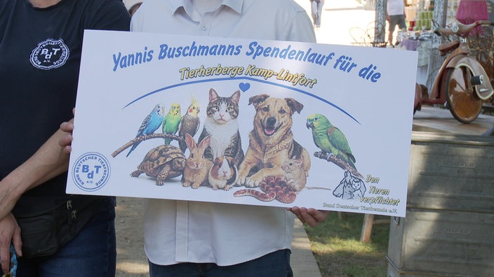 Ein Banner wird gehalten. Darauf sind Haustiere (Hund, Katze, Wellensittiche) sowie die Aufschrift "Yannis Buschmanns Spendenlauf für die Tierherberge Kamp-Lintfort" abgebildet.