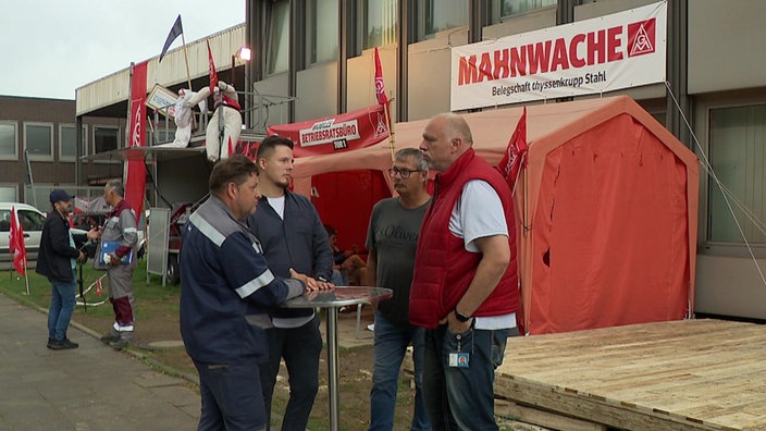 Vier Männer stehen um einen runden Tisch draußen. Hinter ihnnen sind Zelte und Banner mit der Aufschrift "Mahnwache".