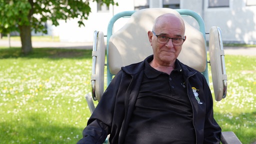 Ein älterer Mann mit Halbglatze und Brille sitzt in einem größeren Transportstuhl auf einer Gänseblümchen-Wiese und lächelt in die Kamera