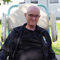 Ein älterer Mann mit Halbglatze und Brille sitzt in einem größeren Transportstuhl auf einer Gänseblümchen-Wiese und lächelt in die Kamera