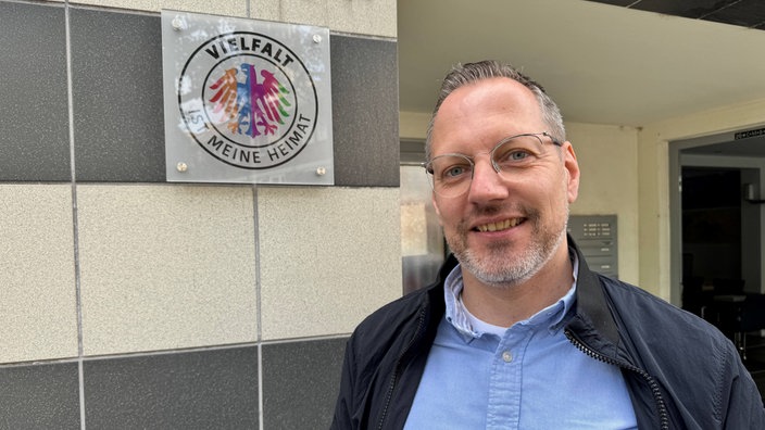 Sebastian Worms lächelt in die Kamera und steht neben einem Schild mit der Aufschtrift "Vielfalt ist meine Heimat".