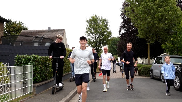 Ein junger Mann im Laufoutfit mit Kappe läuft lächelnd auf der Straße. Hinter ihm laufen weitere Menschen mit ihm auf der Straße. Auf dem Gehweg eine Person auf einem E-Roller.