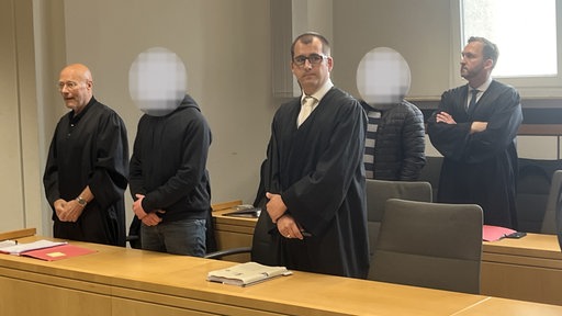 Mehrere Personen stehen in einem Gerichtssaal, einige tragen schwarze Roben, zwei Gesichter sind unkenntlich gemacht.
