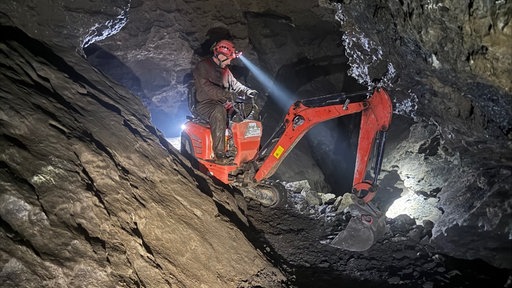 Ein Mann sitzt auf einem kleinen Bagger in einer Höhle und leuchtet mit einer Stirnlampe