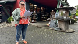Frau hält Dokumente in der Hand. Hinter ihr ist ein Hof mit Scheune.
