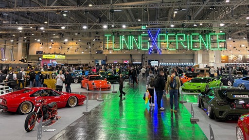 In der großen Messehalle in Essen sind viele verschiedene Autos ausgestellt, Besucherinnen und Besucher schauen sich die Autos an