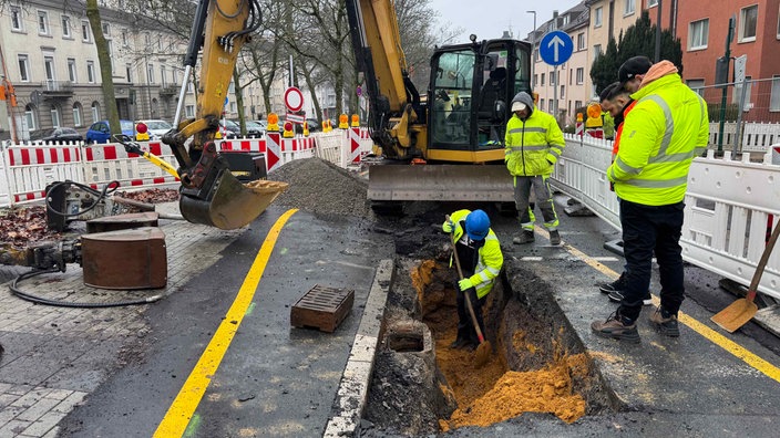 Arbeiter stehen in einem abgesperrten Bereich der Straße vor einem Bagger. In einem Loch steht ein Mann mit einer Schaufel.