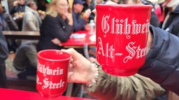 Auf dem Foto sind zwei Hände, die mit Glühweintassen anstoßen. Auf den Tassen steht "Glühwein, Alt-Steele".