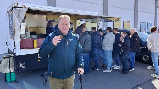 Ein Mann mit Rollator hält eine Frikadelle in der Hand, im Hintergrund ein Imbisswagen, vor dem Menschen Schlange stehen