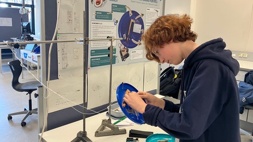 Junge klebt etwas unter eine Frisbee Scheibe an einem Schreibtisch mit Plakat im Hintergrund