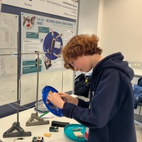 Junge klebt etwas unter eine Frisbee Scheibe an einem Schreibtisch mit Plakat im Hintergrund