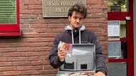 Mann blickt mit ernster Miene in die Kamera. Er hält einen offenen Koffer mit Geld in der Hand und einen zehn Euro Schein in die Höhe.