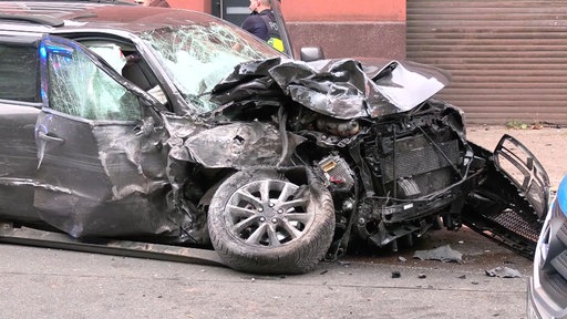 Stark beschädigter schwarzer Pkw mit eingedrückter Front, zerstörter Motorhaube und abgerissenem Vorderrad steht nach einem Unfall am Straßenrand.