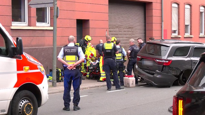 Mehrere Einsatzkräfte von Polizei und Rettungsdienst stehen und knien auf einer Straße neben einem stark beschädigten Pkw.