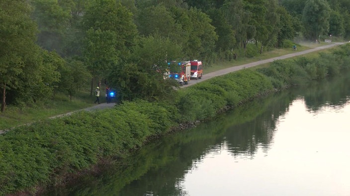 Einsatzwagen der Feuerwehr und Polizei stehen auf dem Weg neben einem Fluss.