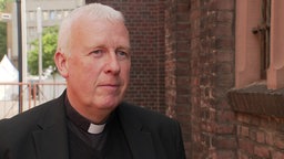 Ein katholischer Pfarrer in schwarzem Hemd und schwarzem Jacket steht vor einer Kirche