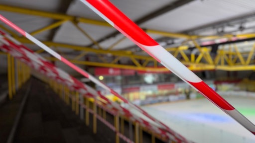 Rot-weißes Absperrband über den gelben Geländern der Tribüne in der Eissporthalle, im Hintergrund die Eisfläche.