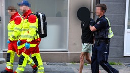 Ein Mann wird von einem Polizeibeamten abgeführt, vor ihnen laufen zwei Sanitäter auf dem Bürgersteig.