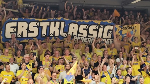 Jubelnde Fans in gelben Trikots, darüber ein Banner mit der Aufschrift "ERSTKLASSIG".