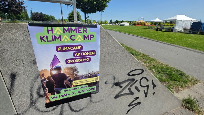Ein buntes Plakat hängt an einer Mauer. Im Hintergrund ist eine Wiese mit aufgebauten Campingzelten zu sehen. 