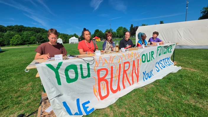 Sechs Personen sitzen draußen auf dem Rasen nebeneinander an einem Tisch, an dem ein Banner mit der Aufschrift "You burn our future. We burn your system" herunterhängt.