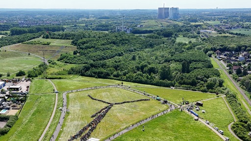 Viele Menschen bilden die Form einer Windkraftanlage, im Hintergrund Kraftwerke
