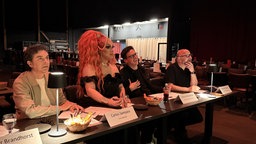 Eine Drag Queen und drei Männer sitzen an einem Tisch und schauen gespannt Richtung Bühne