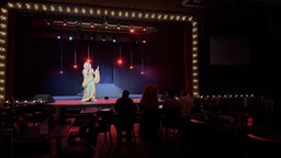 Eine Drag Queen steht auf einer Bühne, davor sitzen vier Personen und schauen Richtung Bühne