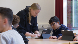 Das Bild zeigt eine Lehrerin, die neben einem Schüler steht. Beide schauen auf ein Tablet