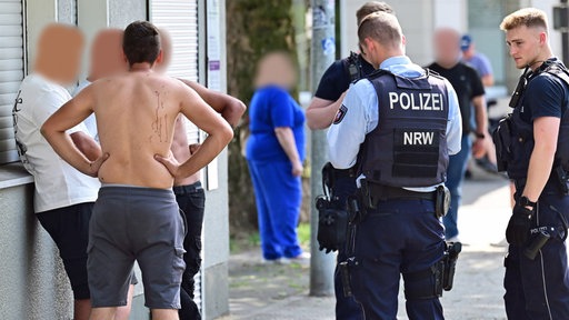 Vier Männer, teils mit nackten Oberkörper, stehen vor einem Haus. Einer von ihnen hat Blutspuren auf dem Rücken. Neben den Männern stehen drei Polizisten.