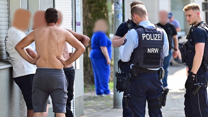 Vier Männer, teils mit nackten Oberkörper, stehen vor einem Haus. Einer von ihnen hat Blutspuren auf dem Rücken. Neben den Männern stehen drei Polizisten.