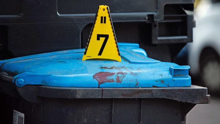 Der Deckel einer Mülltonne ist blutverschmiert. Darauf steht ein Schild der Polizei mit der Nummer 7.