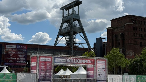 Der ehemalige Förderturm der Zeche Ewald in Herten ragt in den Himmel, im Vordergrund sind Absperrungen und ein Eingang mit Willkommensschild.
