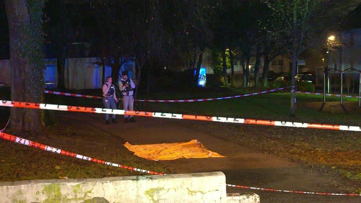 Ein Park bei Nacht. Auf einem Weg liegt ein Tuch, in einen größeren Radius herum iat Polizei Absperrrband, zwei Personen stehen auf dem Weg.