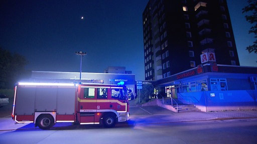 Bei Morgendämmerung steht ein Feuerwehrwagen vor dem Hochhaus. Einsatzkräfte laufen hinein.