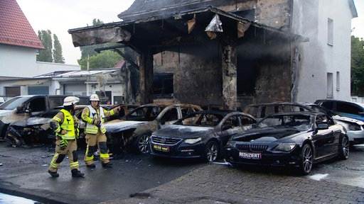 Einsatzkräfte der Feuerwehr laufen vor ausgebrannten Autos entlang. Dahinter befindet sich ein durch den Brand beschädigtes Haus.