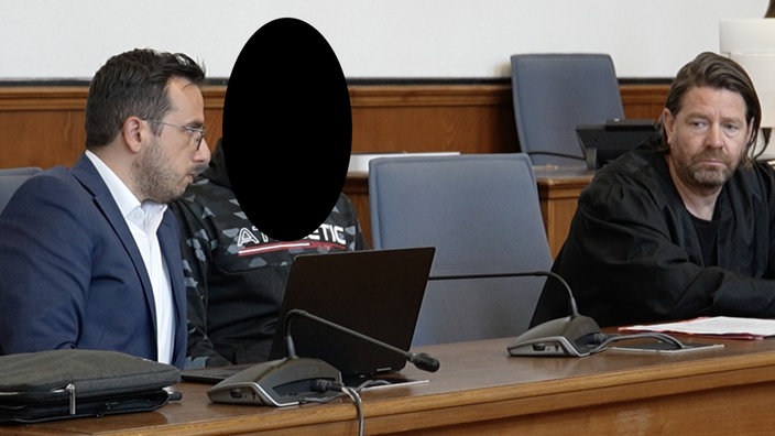 Drei Männer sitzen in einem Gerichtssaal nebeneinander. Der linke spricht mit dem Mann in der Mitte, sein Gesicht ist verpixelt. Rechts sitzt ein Anwalt in seiner Robe. 