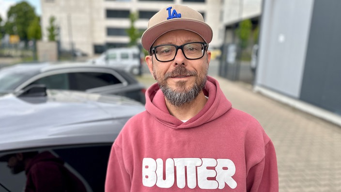 Ein Mann mit einer Kappe steht vor Autos und schaut in die Kamera.