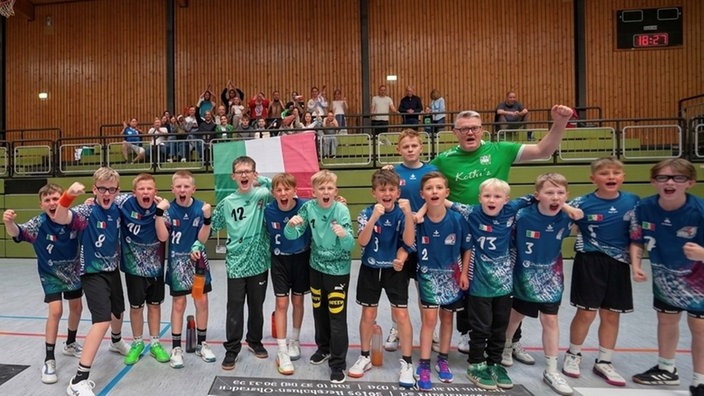 Kinder und Trainer einer Handballmannschaft stehen vor einer gefüllten Tribüne in einer Sporthalle und jubeln.