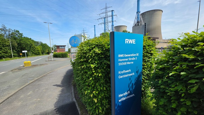 Ein blaues Schild mit der Aufschrift "RWE" steht neben einer Straße, dahinter zwei riesige Kraftwerks-Schornsteine.