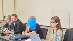 4 Personen im Gerichtssaal. In der Mitte der Angeklagte, der sein Gesicht mit einer blauen Mappe verdeckt. 