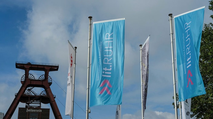 Zwei wehende blaue Fahnen mit der Aufschrift "Lit.Ruhr" stehen neben dem Förderturm der Zeche Zollverein in Essen