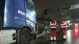 Der Lkw ist auf Schutzplanke aufgefahren, zwei Männer in orangener Arbeitskleidung stehen davor und ein Kran hebt die Wand aus der Fahrbahn