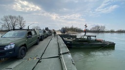 Mehrere Militärfahrzeuge parken an einem Steg, an dem ein Militärboot liegt.