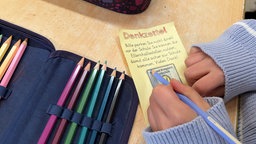 Ein gelber Zettel der ausgemalt wird. Auf diesen steht "Denkzettel" und "Bitte parken Sie nicht direkt vor der Schule. Sie können die Elternhaltestelle nutzen, damit alle sicher zur Schule kommen. Vielen Dank!"