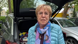 Birgit Westphal mit Brille, hellblauer Jacke und buntem Schal steht vor einem offenen Kofferraum, im Hintergrund sind Autos und Bäume.