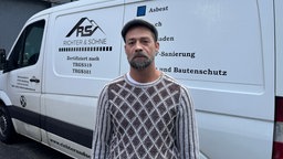 David Richter mit grauem Bart, gemustertem Pullover und Kappe steht vor einem Firmenfahrzeug.