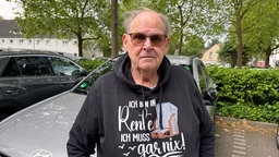 Rentner Hans Kirchberg steht im Freien vor geparkten Autos und Bäumen.