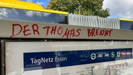 Es ist ein Fahrplanschaukasten an einer Bushaltestelle in Essen zu sehen auf dessen oberen Rahmen in roter Druckschrift in Großbuchstaben "Der Thomas brennt" geschrieben wurde.