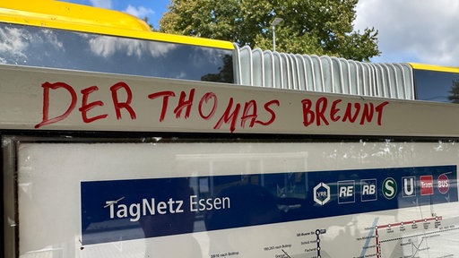 Es ist ein Fahrplanschaukasten an einer Bushaltestelle in Essen zu sehen auf dessen oberen Rahmen in roter Druckschrift in Großbuchstaben "Der Thomas brennt" geschrieben wurde.